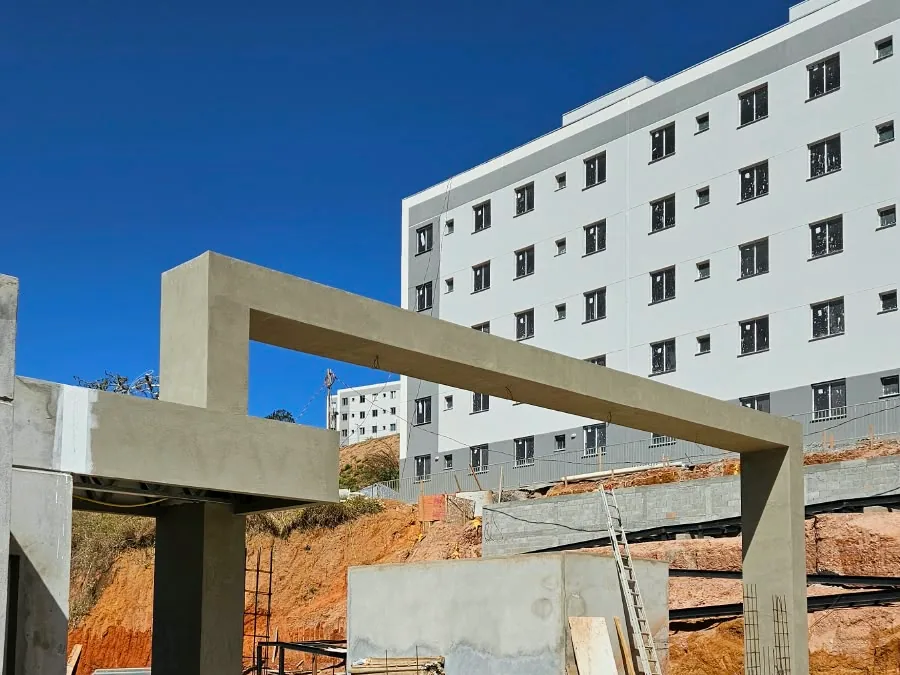 Pórtico em obra com acabamento liso cinza-areia, uma coluna de apoio e um prédio residencial branco ao fundo, num canteiro com terra e escoras
