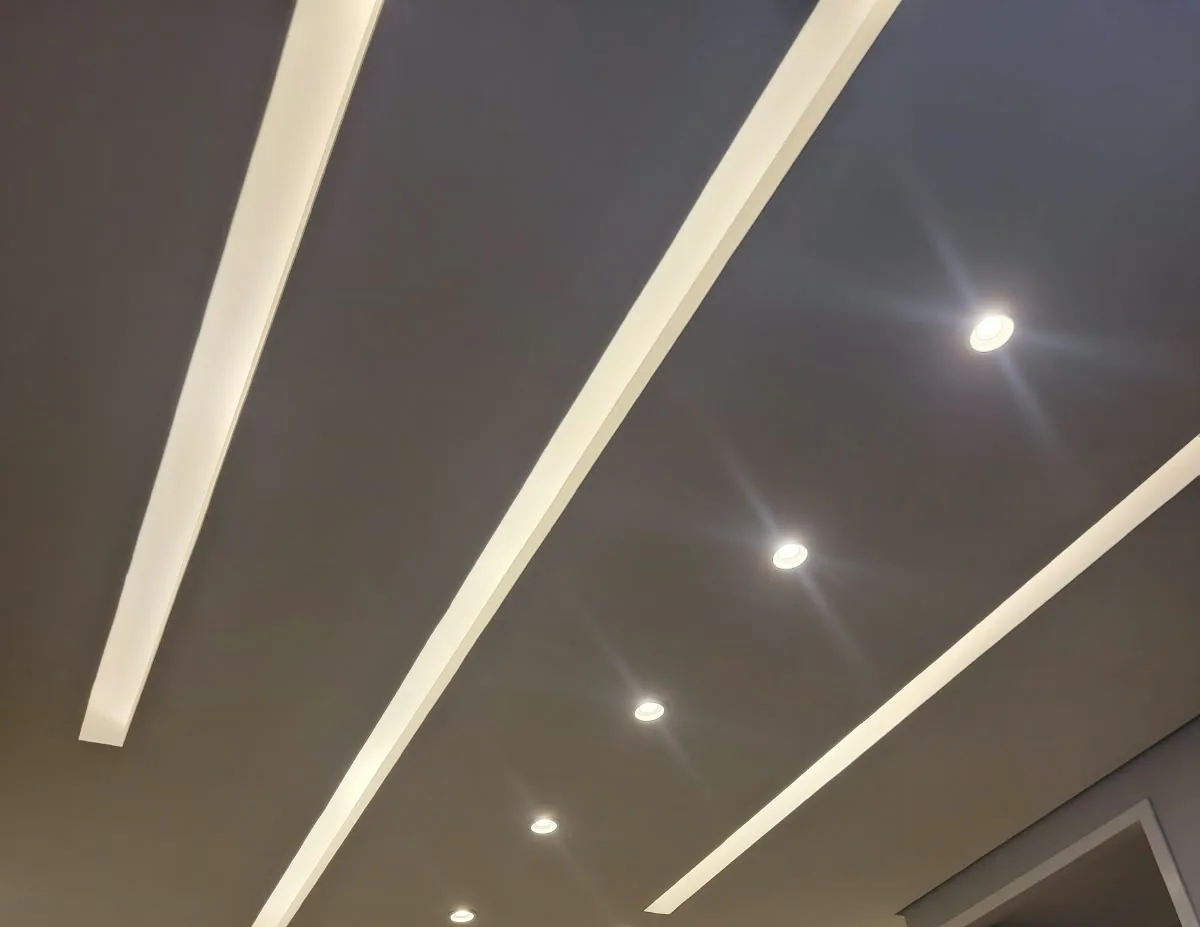 Forro de gesso cinza-escuro com rasgos de luz acesos em linhas retas e spots redondos embutidos, num corredor com portas brancas
