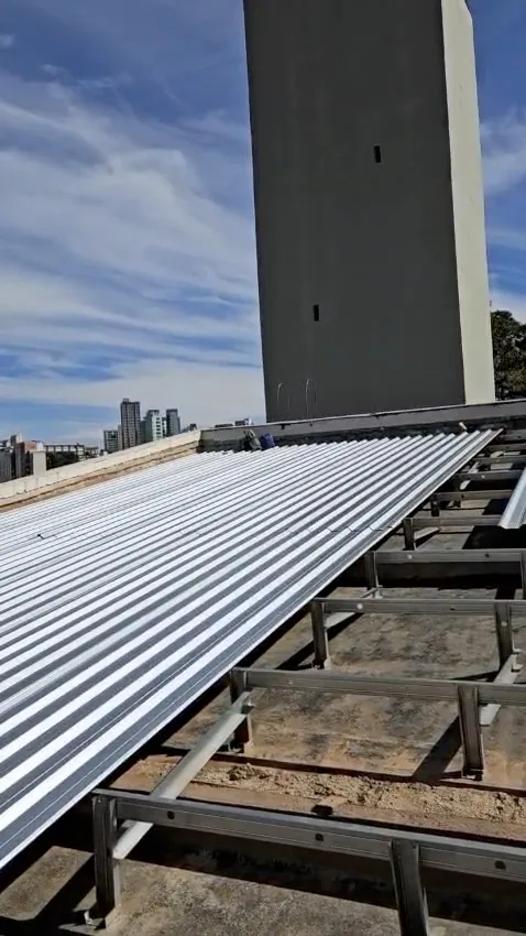 Quadro do vídeo: telhas metálicas já instaladas sobre a estrutura do telhado, com a torre de concreto e a cidade ao fundo